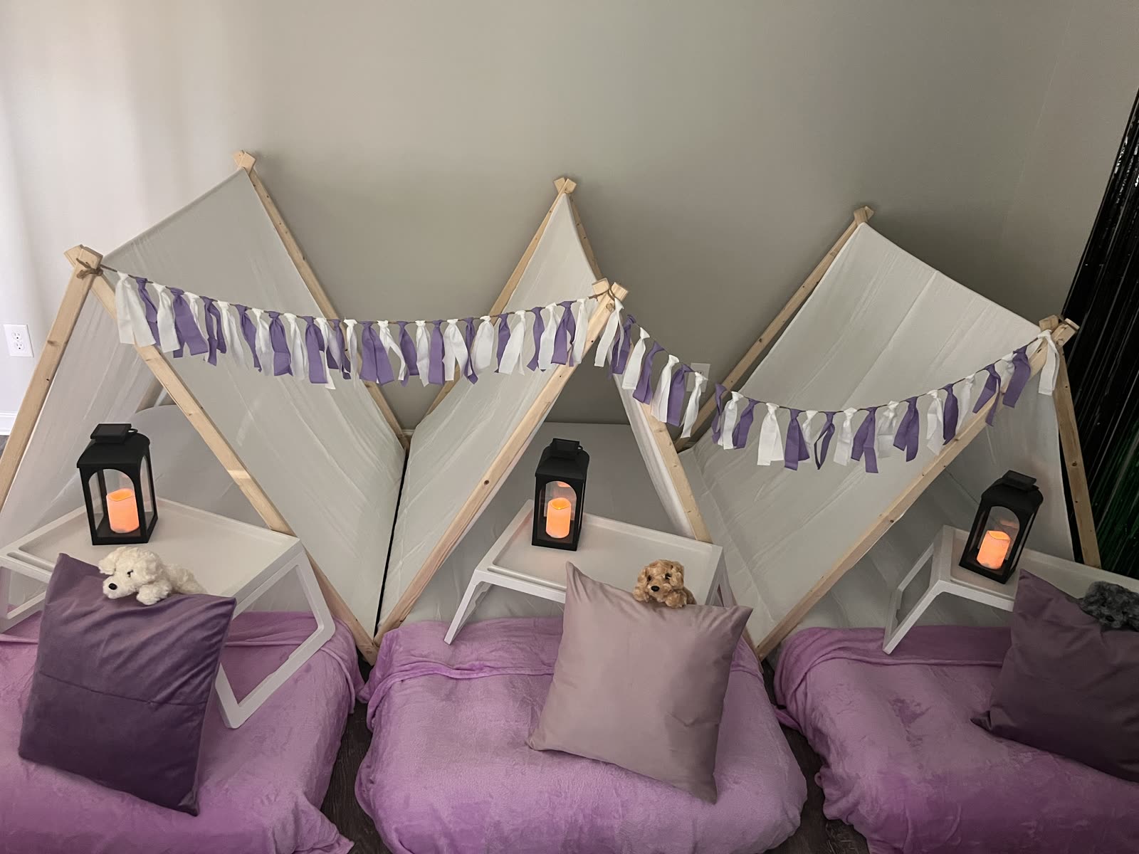 Three white canvas teepee tents with purple and white tassel garland and lit lanterns, set up in a living room
