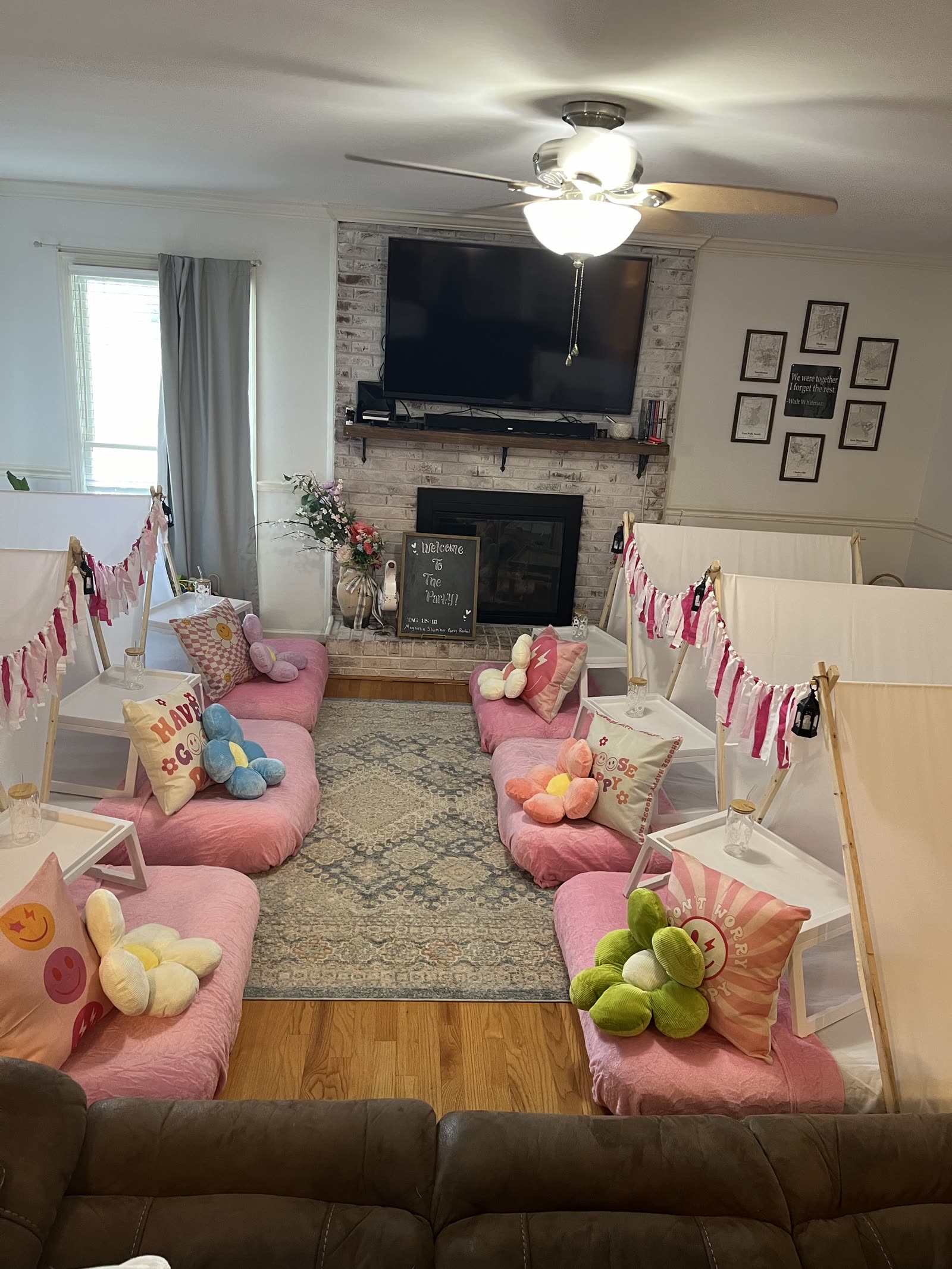 Six pink teepee sleepover beds arranged around a living room rug with a welcome chalkboard at the fireplace